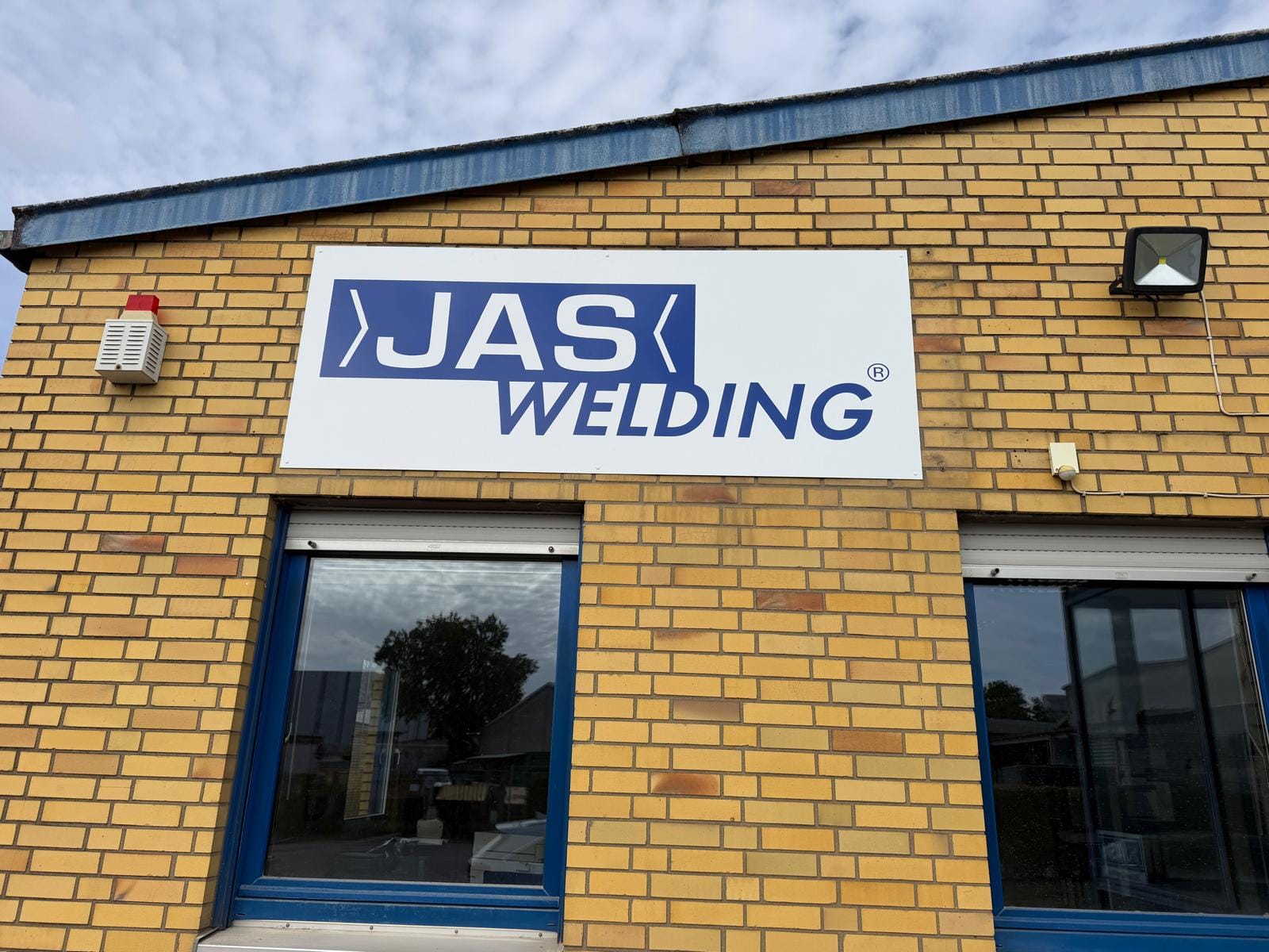 Weißes Firmenschild mit blauem Logo JAS Welding an einer gelben Klinkerfassade